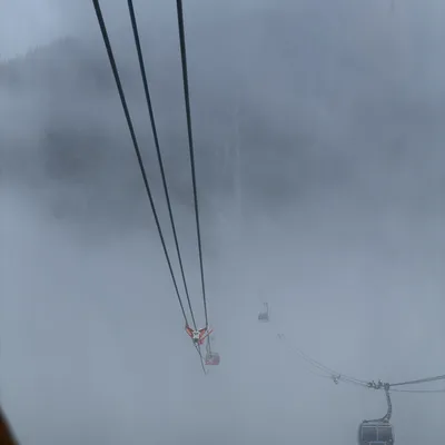 This high-angle shot captures cable cars traveling along suspension lines through a dense, foggy mountain landscape. The thick mist obscures the surrounding forest and terrain, creating a dramatic, atmospheric view of the transit system.