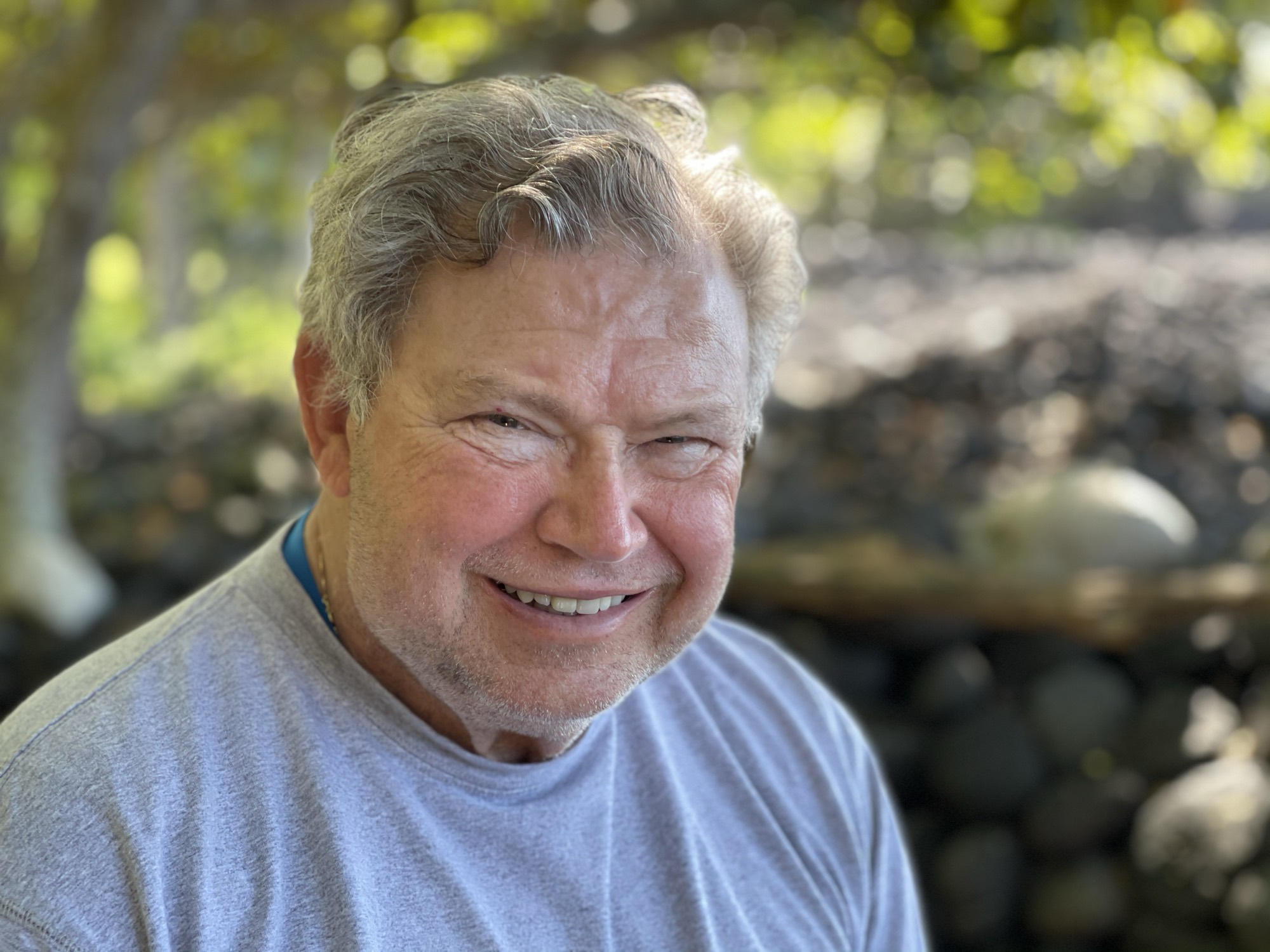 A close-up portrait shows a smiling, older man with grey hair and a light beard wearing a grey t-shirt. He is outdoors with a blurred background of green foliage and dark rocks.