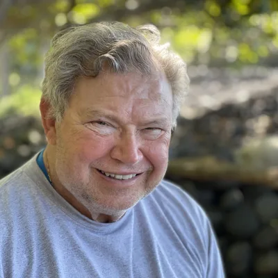 A close-up portrait shows a smiling, older man with grey hair and a light beard wearing a grey t-shirt. He is outdoors with a blurred background of green foliage and dark rocks.