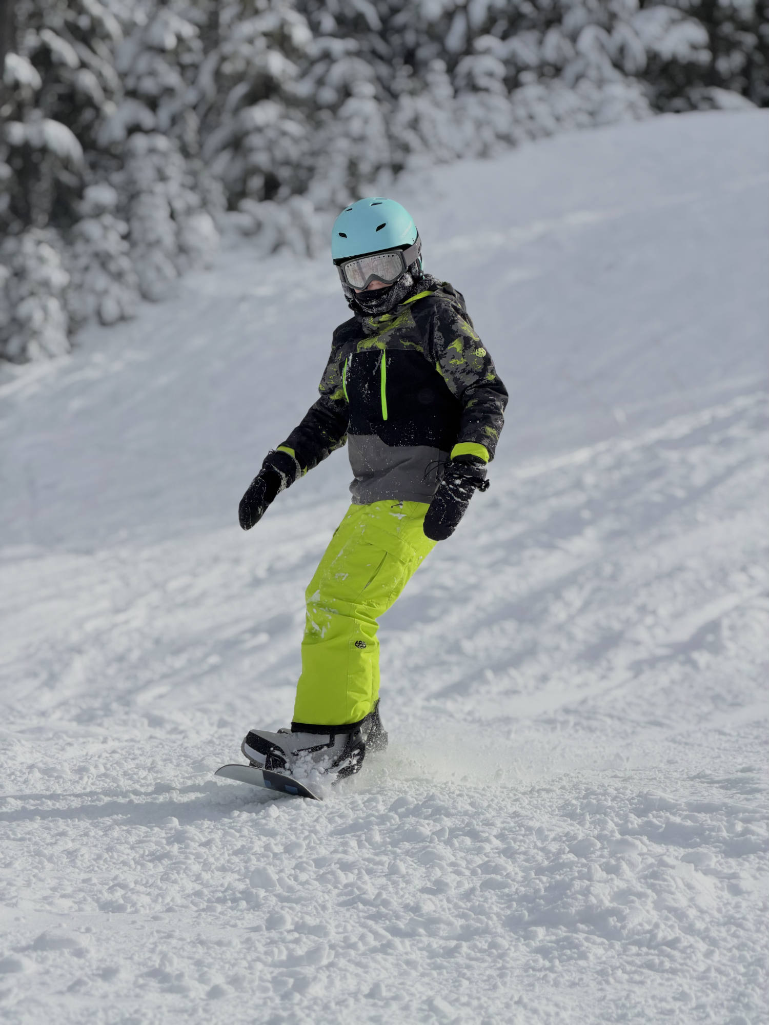 A person wearing a light blue helmet, goggles, and bright yellow-green pants is snowboarding down a snowy slope. The background features a snow-covered hill with pine trees dusted in snow.