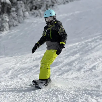 A person wearing a light blue helmet, goggles, and bright yellow-green pants is snowboarding down a snowy slope. The background features a snow-covered hill with pine trees dusted in snow.
