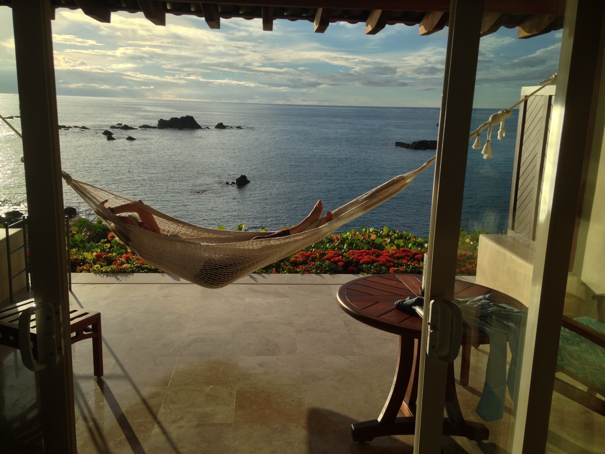 A person relaxes in a woven hammock strung across a tiled patio, which overlooks a calm ocean with rocky outcrops under a cloudy sky. The scene is framed by a sliding glass doorway, with a wooden table and chairs visible on the patio.