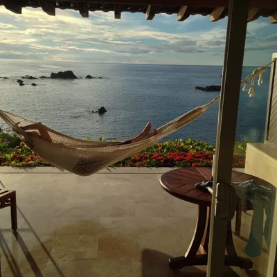A person relaxes in a woven hammock strung across a tiled patio, which overlooks a calm ocean with rocky outcrops under a cloudy sky. The scene is framed by a sliding glass doorway, with a wooden table and chairs visible on the patio.