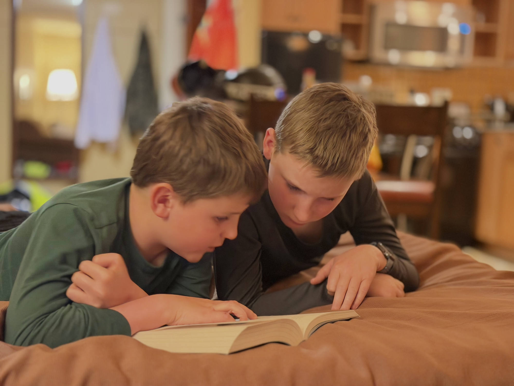 Two young boys lie on their stomachs on a brown bed or cushion, intently reading an open book together. The scene is set indoors, with a softly blurred kitchen and living area visible in the background.