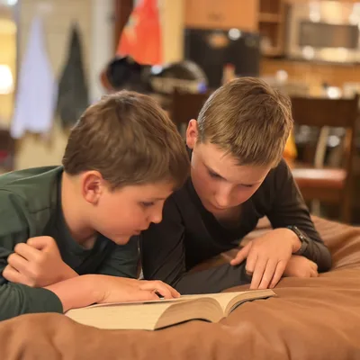 Two young boys lie on their stomachs on a brown bed or cushion, intently reading an open book together. The scene is set indoors, with a softly blurred kitchen and living area visible in the background.