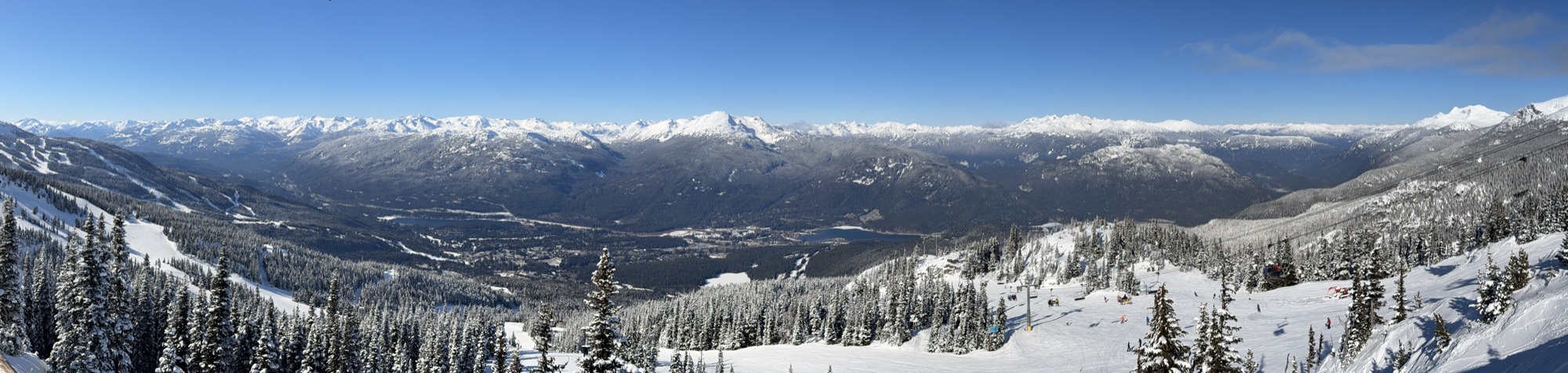 This panoramic photograph captures a wide, scenic view of a snow-covered mountain range and valley on a clear, sunny day. In the foreground, skiers and a chairlift are visible on the snowy slopes of a ski resort, surrounded by snow-dusted evergreen trees.