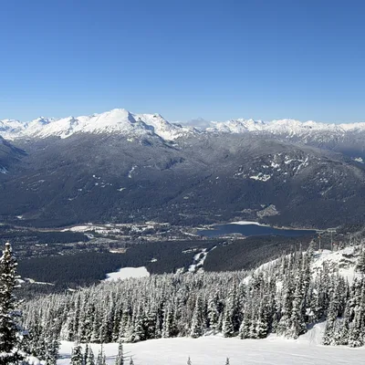 This panoramic photograph captures a wide, scenic view of a snow-covered mountain range and valley on a clear, sunny day. In the foreground, skiers and a chairlift are visible on the snowy slopes of a ski resort, surrounded by snow-dusted evergreen trees.