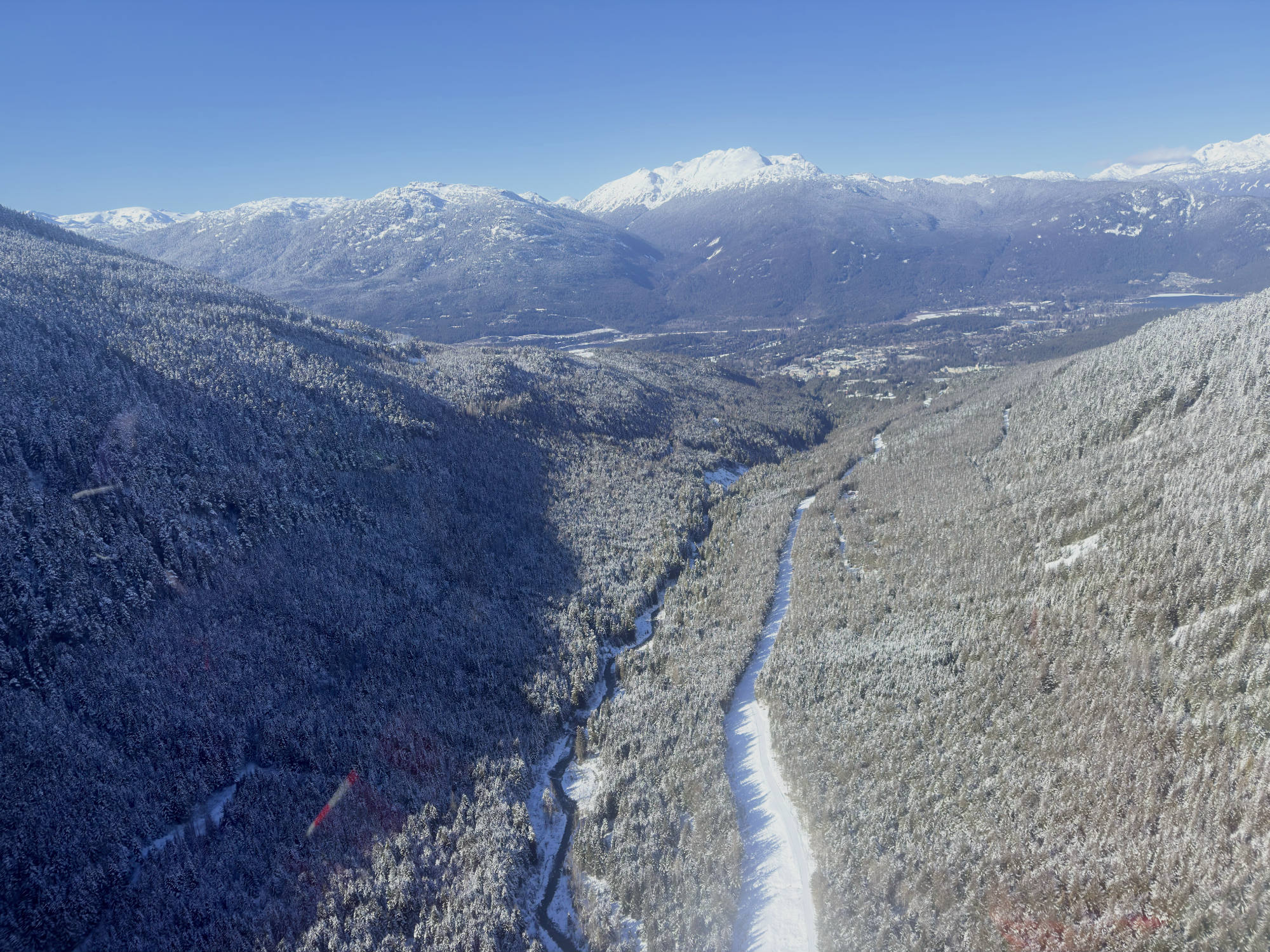 An aerial view captures a snow-covered mountain valley with a winding river and a snow-dusted road cutting through a dense evergreen forest. In the background, majestic, snow-capped mountain peaks rise against a clear blue sky.