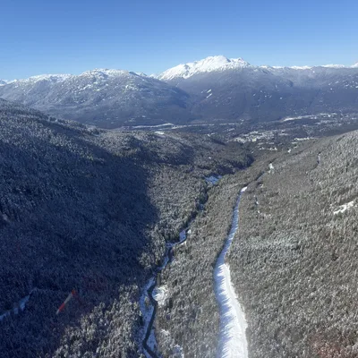 An aerial view captures a snow-covered mountain valley with a winding river and a snow-dusted road cutting through a dense evergreen forest. In the background, majestic, snow-capped mountain peaks rise against a clear blue sky.