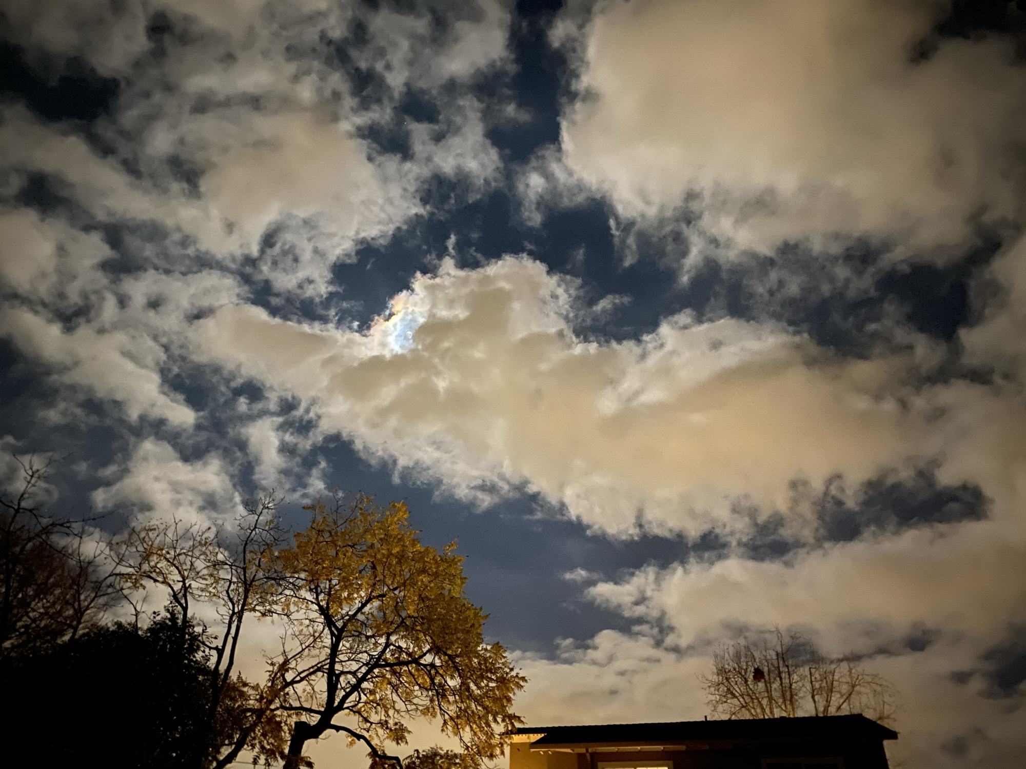 This low-angle shot captures a night sky filled with dramatic, illuminated clouds, with a hint of rainbow-colored iridescence visible where the moon is partially obscured. Below, the silhouettes of trees, including one with yellow autumn leaves, and the roofline of a building frame the bottom of the scene.