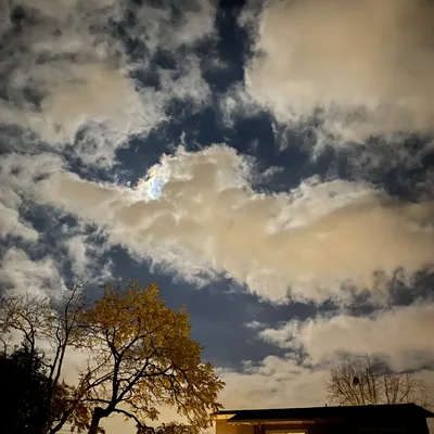 This low-angle shot captures a night sky filled with dramatic, illuminated clouds, with a hint of rainbow-colored iridescence visible where the moon is partially obscured. Below, the silhouettes of trees, including one with yellow autumn leaves, and the roofline of a building frame the bottom of the scene.