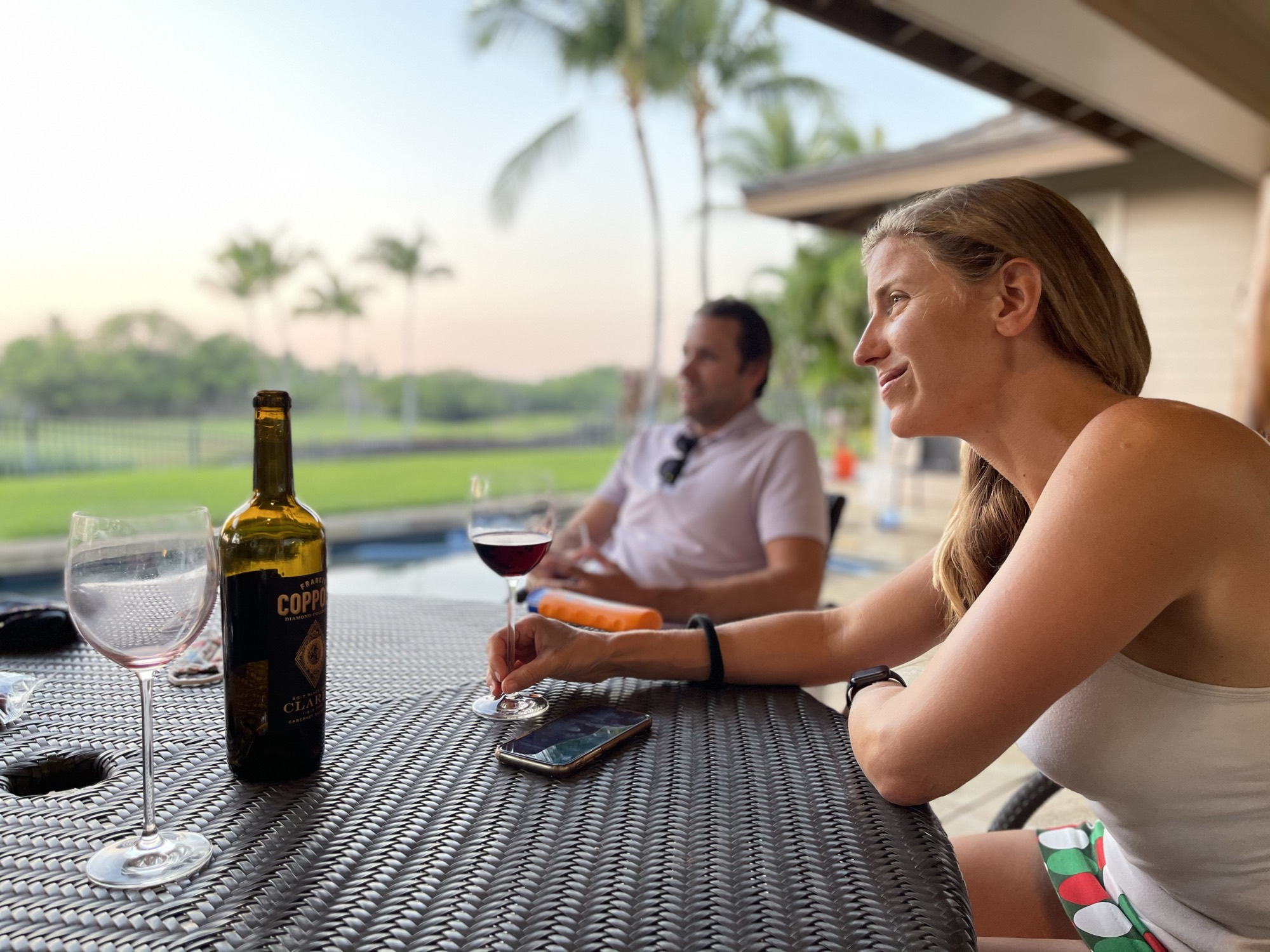 A woman sits at an outdoor wicker table holding a glass of red wine next to a bottle of Coppola wine, while a man sits in the background near a swimming pool and palm trees. They appear to be relaxing on a patio during the evening.