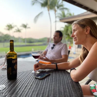 A woman sits at an outdoor wicker table holding a glass of red wine next to a bottle of Coppola wine, while a man sits in the background near a swimming pool and palm trees. They appear to be relaxing on a patio during the evening.