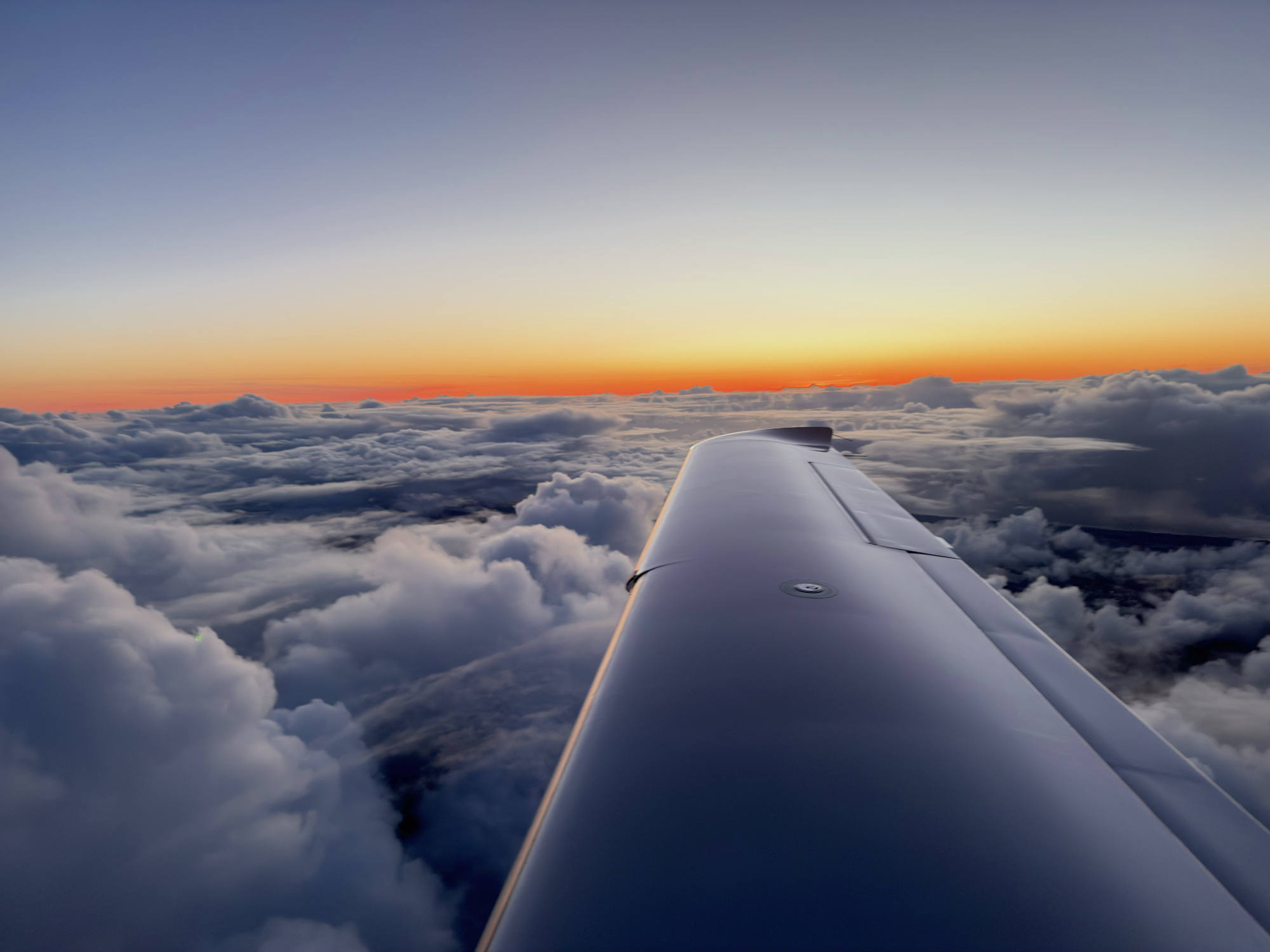 An aerial view from an airplane window captures the aircraft's wing extending over a dense layer of clouds during a vibrant sunset. The horizon glows with bright orange and yellow light beneath a clear, fading blue sky.
