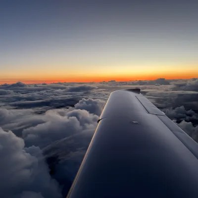 An aerial view from an airplane window captures the aircraft's wing extending over a dense layer of clouds during a vibrant sunset. The horizon glows with bright orange and yellow light beneath a clear, fading blue sky.