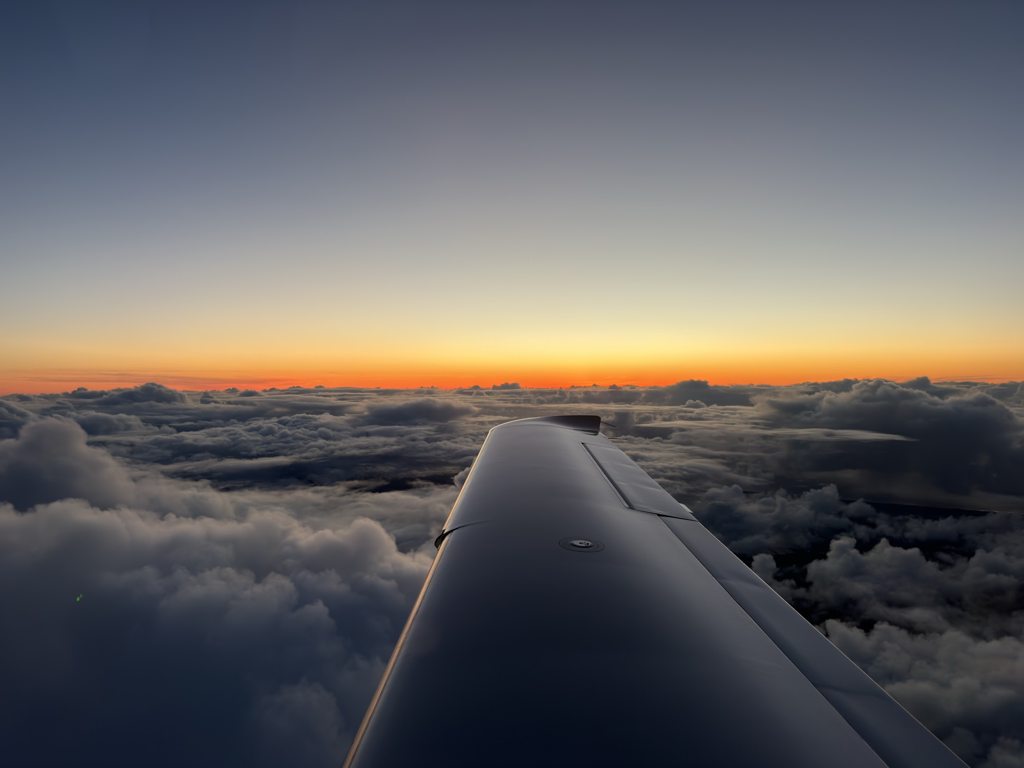 An aerial view from an airplane window shows the aircraft's wing extending over a dense layer of clouds. In the distance, a vibrant orange and yellow sunset glows along the horizon beneath a clear, fading blue sky.