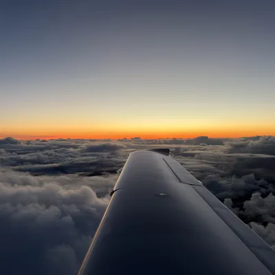 An aerial view from an airplane window shows the aircraft's wing extending over a dense layer of clouds. In the distance, a vibrant orange and yellow sunset glows along the horizon beneath a clear, fading blue sky.