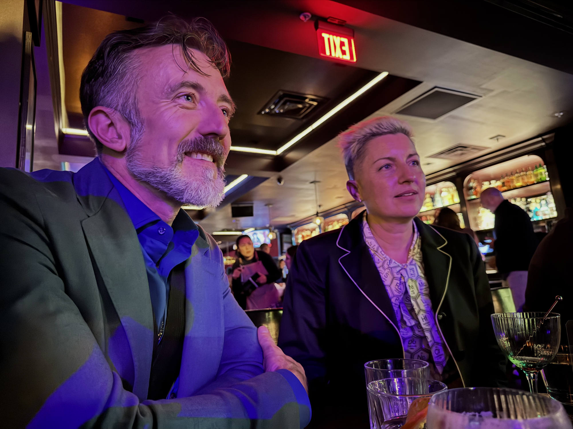 A man with a beard and a woman with short blonde hair sit together at a bar, looking off-camera with smiles. The dimly lit venue features colorful ambient lighting, an illuminated exit sign, and other patrons in the background.