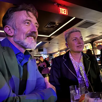 A man with a beard and a woman with short blonde hair sit together at a bar, looking off-camera with smiles. The dimly lit venue features colorful ambient lighting, an illuminated exit sign, and other patrons in the background.