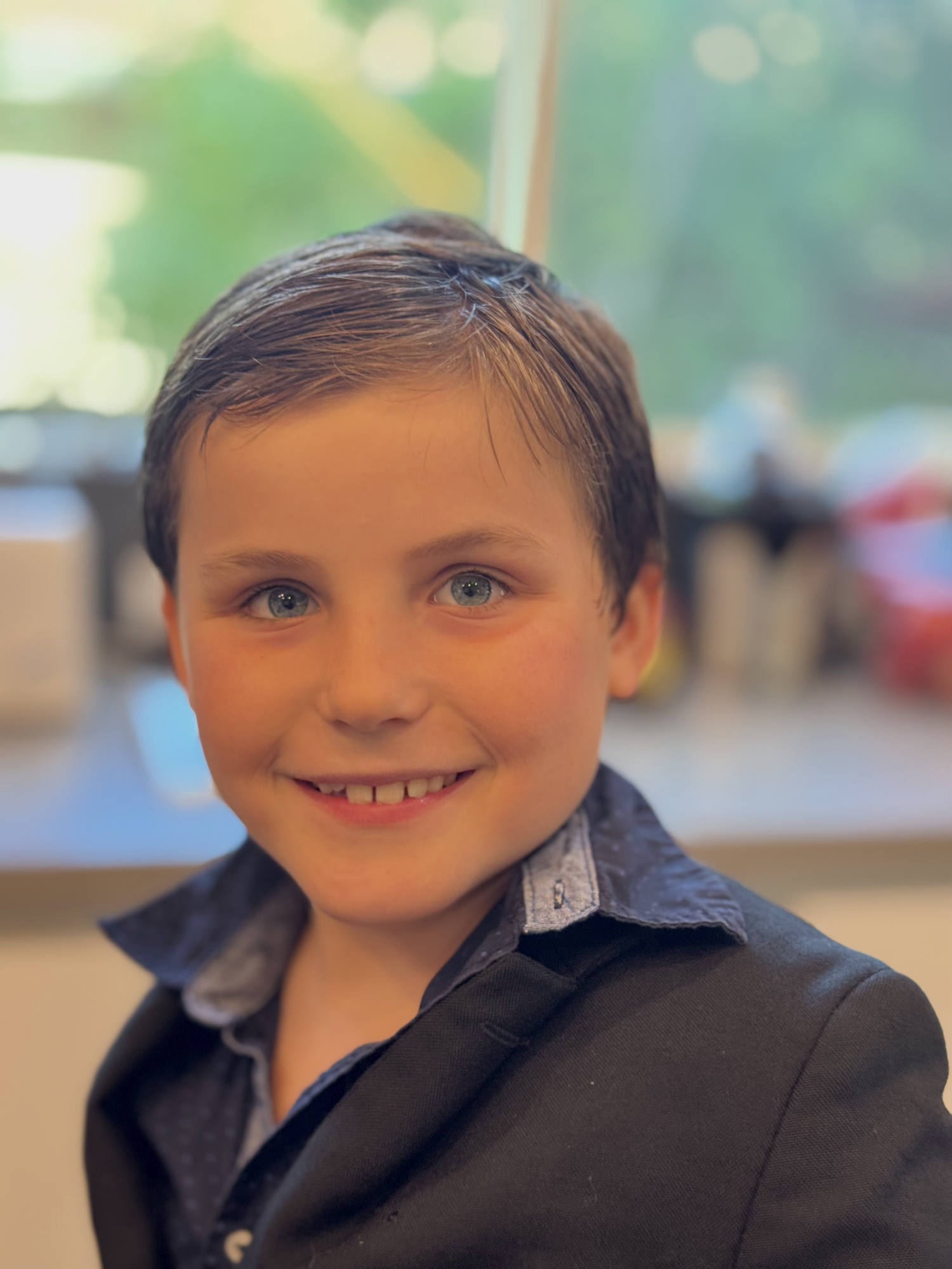 A close-up portrait of a young boy with blue eyes smiling at the camera while wearing a dark jacket over a collared shirt. He is indoors, with a softly blurred background showing a window and hints of an interior room.