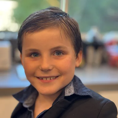 A close-up portrait of a young boy with blue eyes smiling at the camera while wearing a dark jacket over a collared shirt. He is indoors, with a softly blurred background showing a window and hints of an interior room.