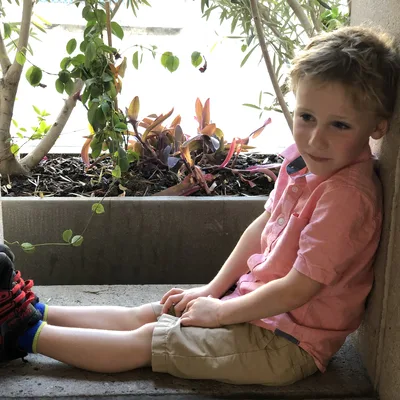 A young boy in a pink shirt and khaki shorts sits in a concrete alcove with his legs stretched out and his back against the wall. Behind him, a planter box contains green and reddish-purple plants.