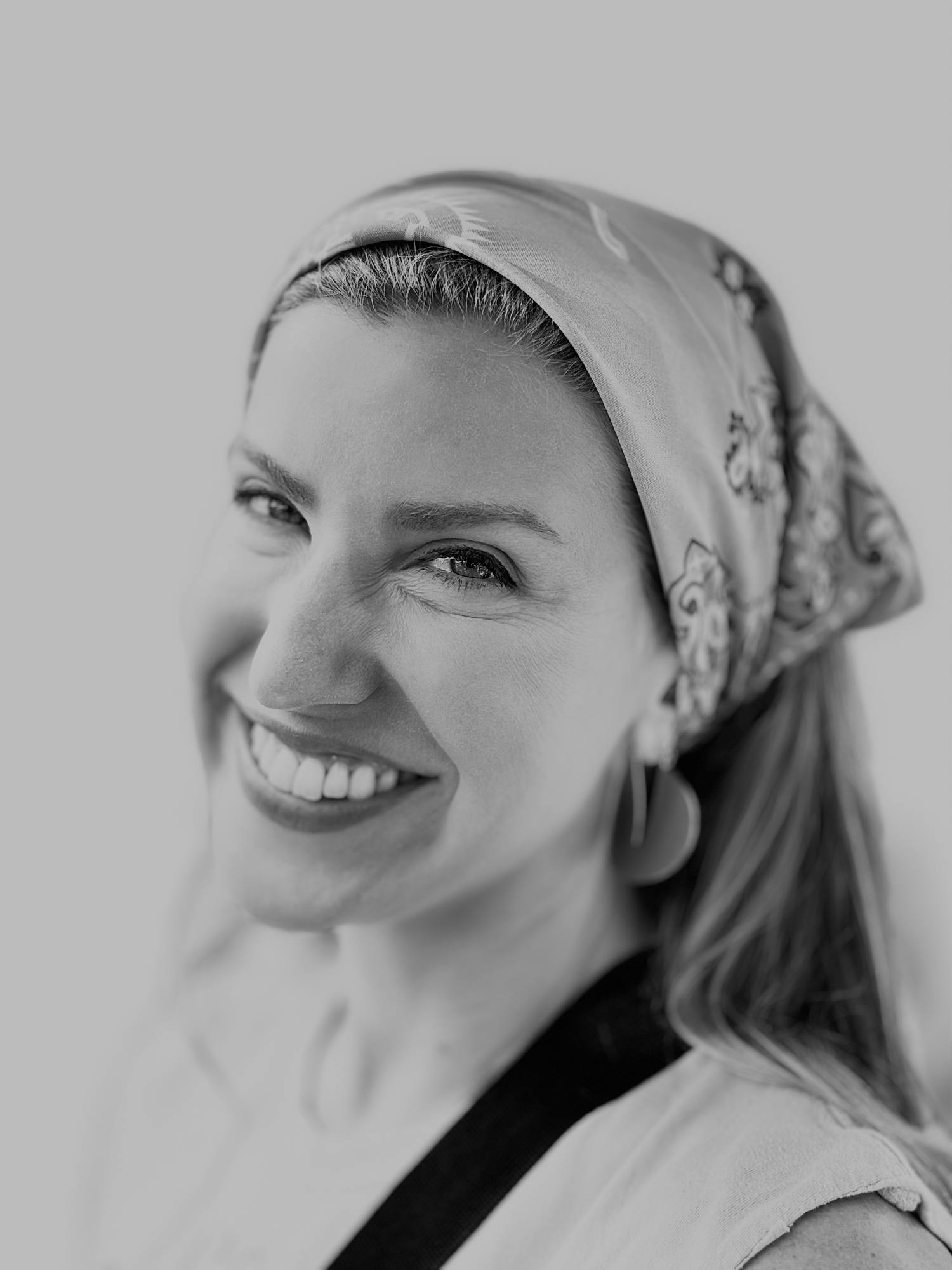 A black and white, close-up portrait of a smiling woman wearing a patterned headscarf and large earrings. She is looking directly at the camera against a plain, bright background.