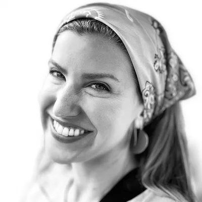 A black and white, close-up portrait of a smiling woman wearing a patterned headscarf and large earrings. She is looking directly at the camera against a plain, bright background.
