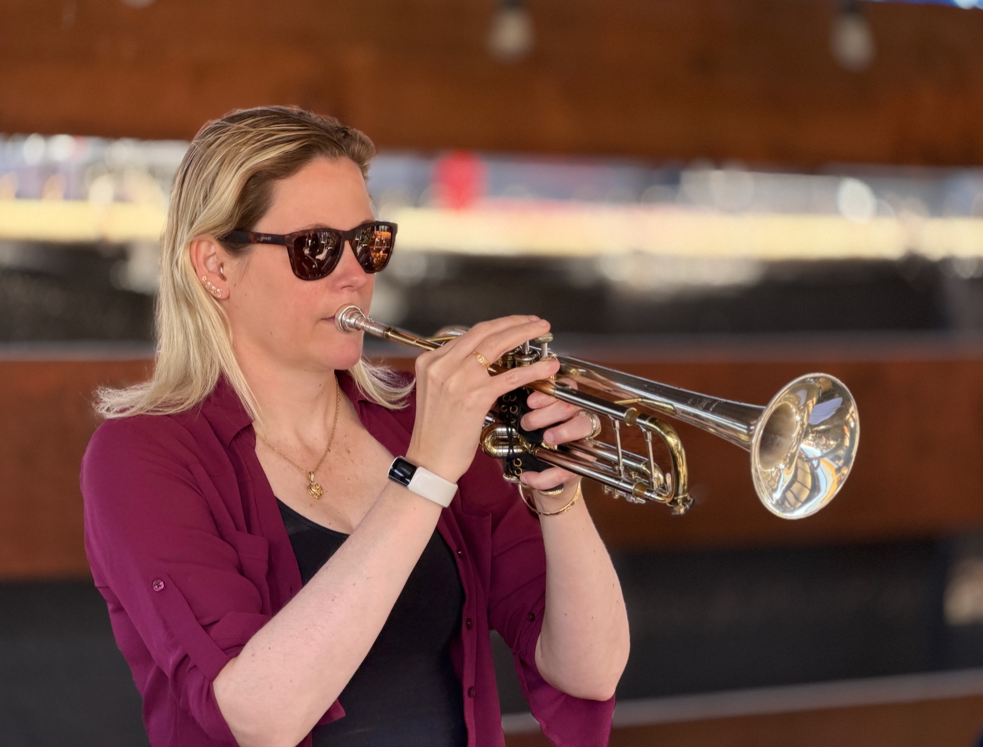 A blonde woman wearing sunglasses and a maroon shirt plays a silver and gold trumpet. She is captured in a medium close-up shot against a softly blurred, warm-toned background.