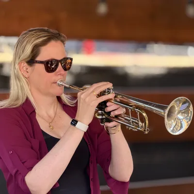 A blonde woman wearing sunglasses and a maroon shirt plays a silver and gold trumpet. She is captured in a medium close-up shot against a softly blurred, warm-toned background.