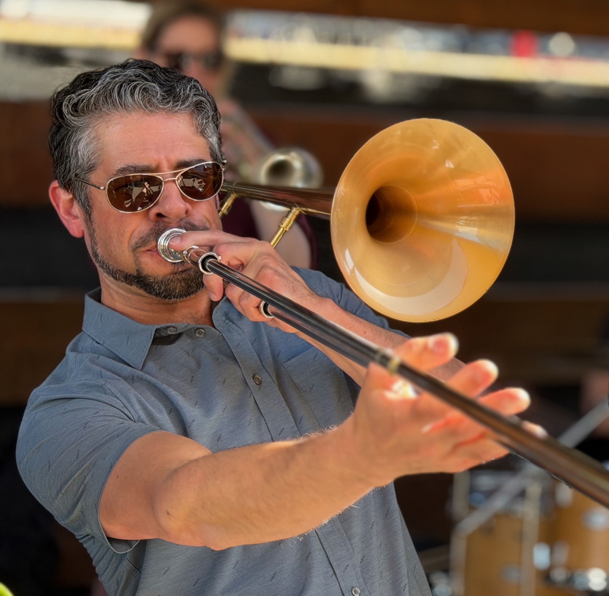 A close-up shot captures a man with sunglasses playing a brass trombone outdoors, with another musician blurred in the background. He is wearing a grey short-sleeved button-down shirt and extending the trombone's slide forward.
