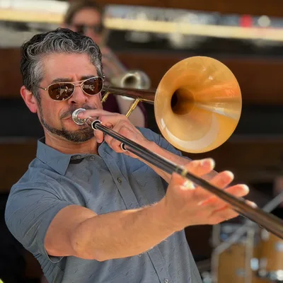 A close-up shot captures a man with sunglasses playing a brass trombone outdoors, with another musician blurred in the background. He is wearing a grey short-sleeved button-down shirt and extending the trombone's slide forward.