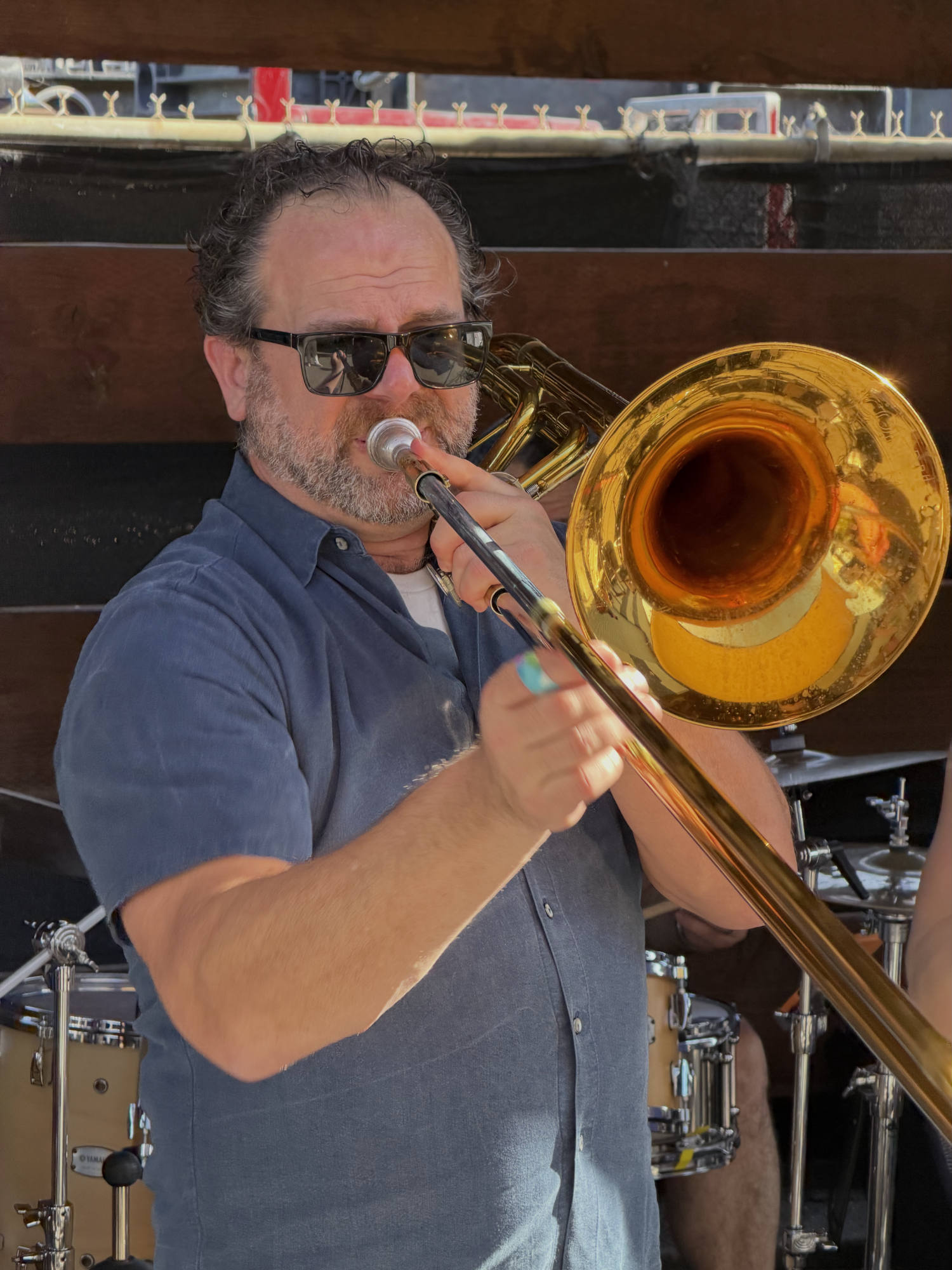 A man wearing sunglasses and a blue button-down shirt plays a brass trombone on an outdoor stage. A drum kit is visible in the background behind him.