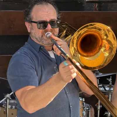 A man wearing sunglasses and a blue button-down shirt plays a brass trombone on an outdoor stage. A drum kit is visible in the background behind him.