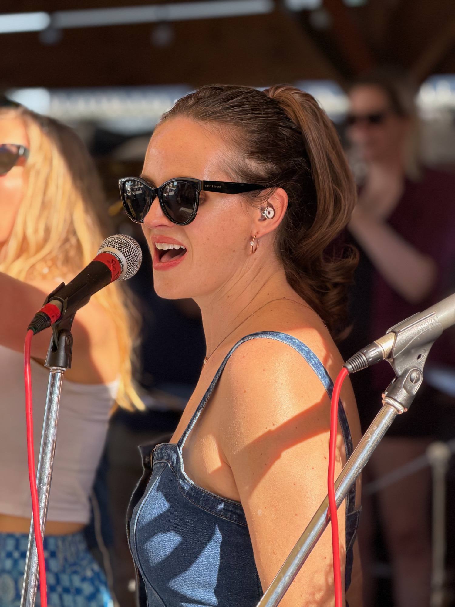 A woman wearing sunglasses, a denim top, and an in-ear monitor sings into a microphone during an outdoor performance. Other band members and equipment are visible in the background of the sunny stage setting.
