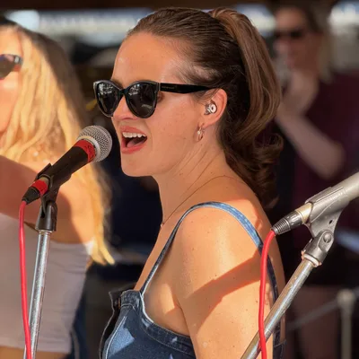 A woman wearing sunglasses, a denim top, and an in-ear monitor sings into a microphone during an outdoor performance. Other band members and equipment are visible in the background of the sunny stage setting.
