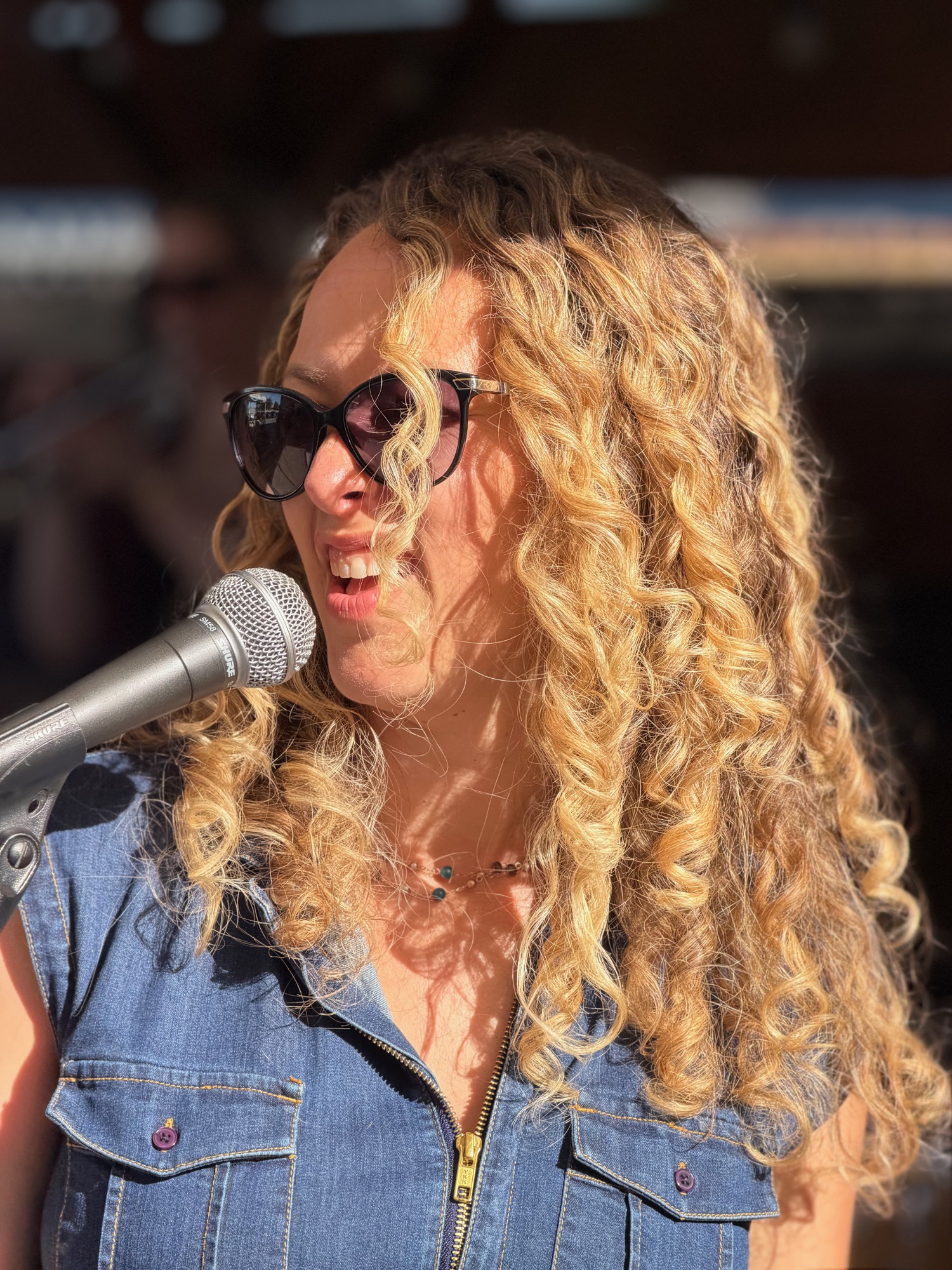 A close-up shot shows a woman with long, curly blonde hair and sunglasses smiling as she sings or speaks into a microphone. She is wearing a denim shirt and is positioned outdoors in bright sunlight, with a blurred background suggesting a stage or performance setting.