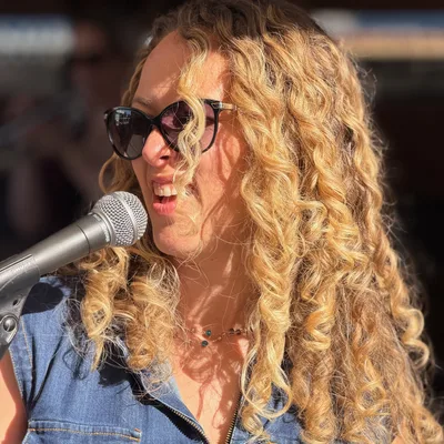 A close-up shot shows a woman with long, curly blonde hair and sunglasses smiling as she sings or speaks into a microphone. She is wearing a denim shirt and is positioned outdoors in bright sunlight, with a blurred background suggesting a stage or performance setting.