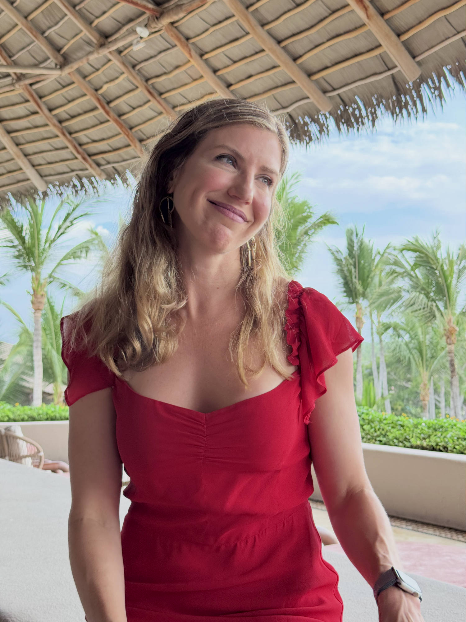 A smiling woman in a red dress sits under a thatched roof structure, looking off to the side. The background features palm trees and a blue sky, suggesting a tropical resort or vacation setting.