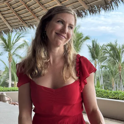 A smiling woman in a red dress sits under a thatched roof structure, looking off to the side. The background features palm trees and a blue sky, suggesting a tropical resort or vacation setting.