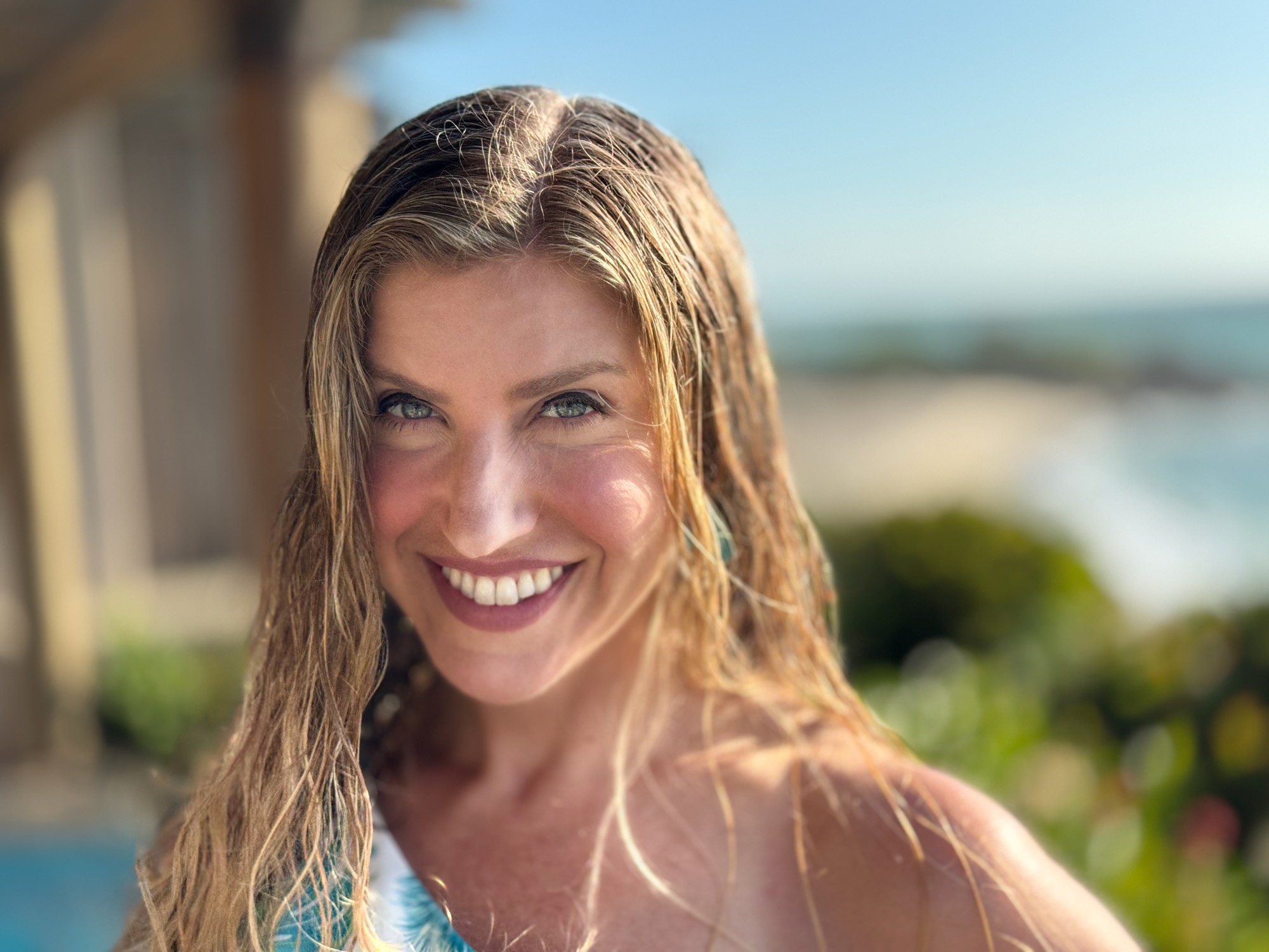 A close-up portrait shows a smiling woman with wet blonde hair looking directly at the camera. She is outdoors on a sunny day, with a blurred background of a beach, ocean, and coastal vegetation.