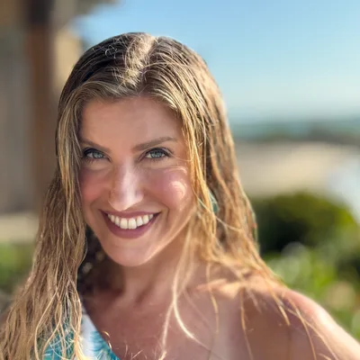 A close-up portrait shows a smiling woman with wet blonde hair looking directly at the camera. She is outdoors on a sunny day, with a blurred background of a beach, ocean, and coastal vegetation.
