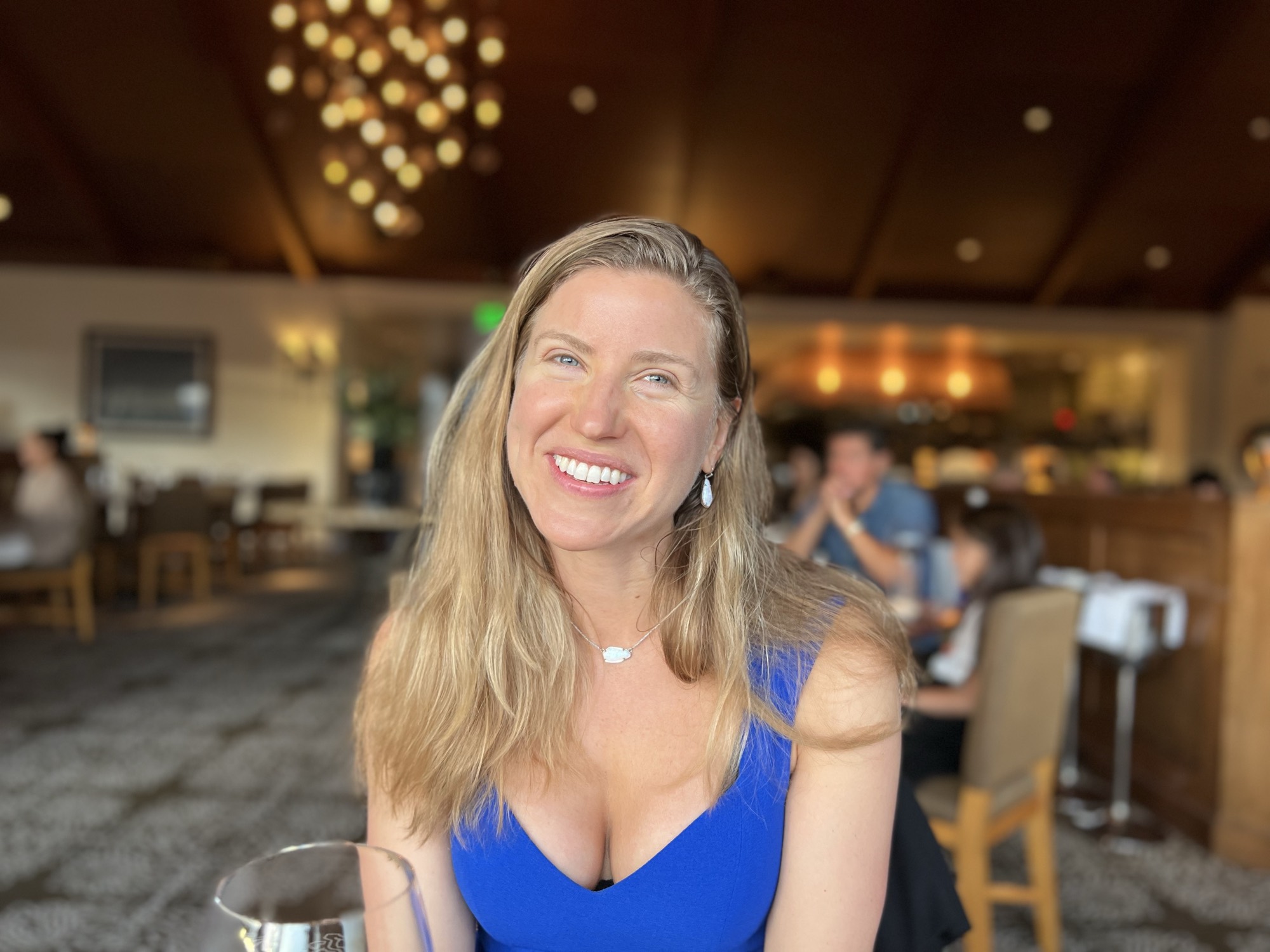 A smiling woman with blonde hair and a blue dress sits at a table in a restaurant, with a wine glass partially visible in the foreground. The background shows other patrons and the warm lighting of the dining room.