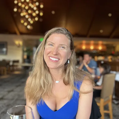 A smiling woman with blonde hair and a blue dress sits at a table in a restaurant, with a wine glass partially visible in the foreground. The background shows other patrons and the warm lighting of the dining room.