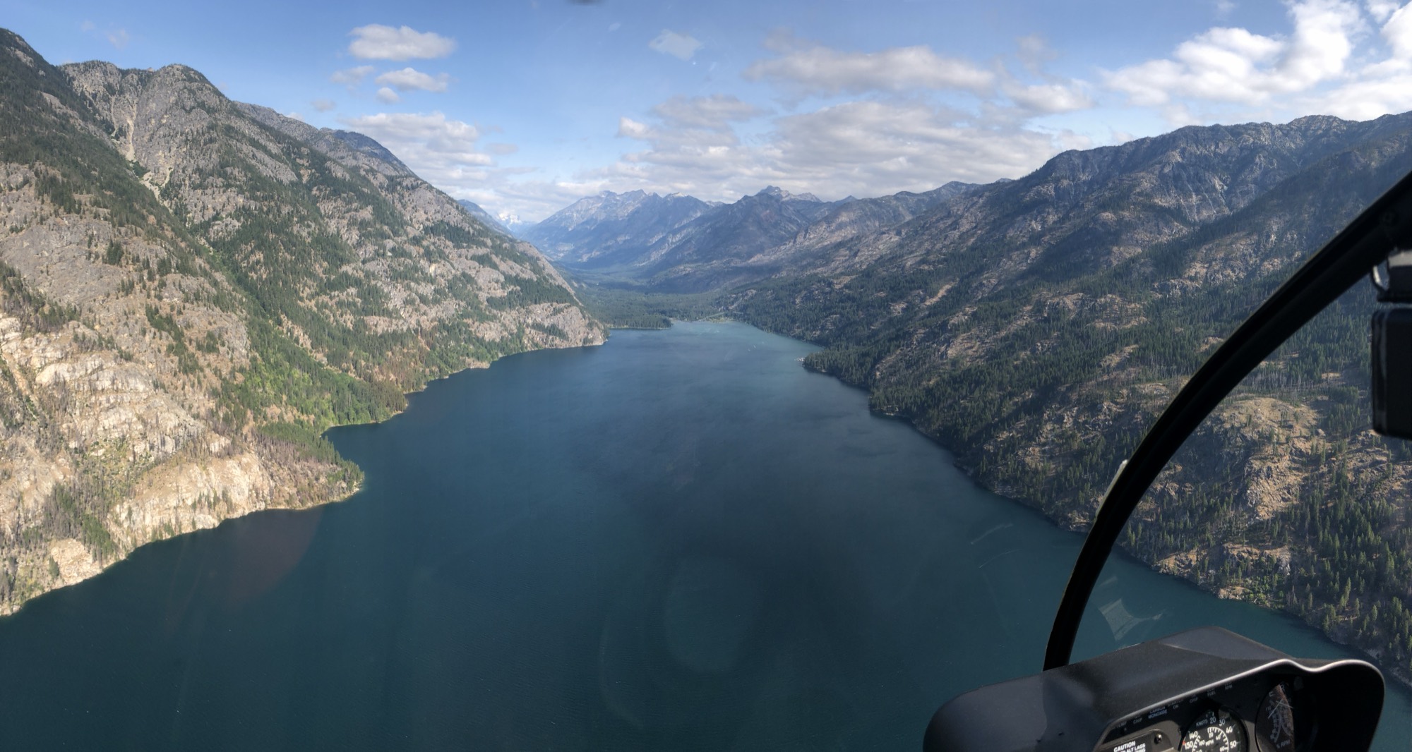 An aerial view from inside a helicopter cockpit shows a long, deep blue lake winding through a steep mountain valley under a partly cloudy sky. The forested, rocky slopes of the mountains rise sharply on both sides of the water, with the helicopter's instrument panel and window frame visible in the bottom right corner.