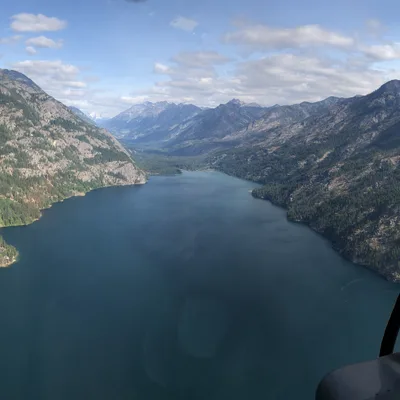 An aerial view from inside a helicopter cockpit shows a long, deep blue lake winding through a steep mountain valley under a partly cloudy sky. The forested, rocky slopes of the mountains rise sharply on both sides of the water, with the helicopter's instrument panel and window frame visible in the bottom right corner.
