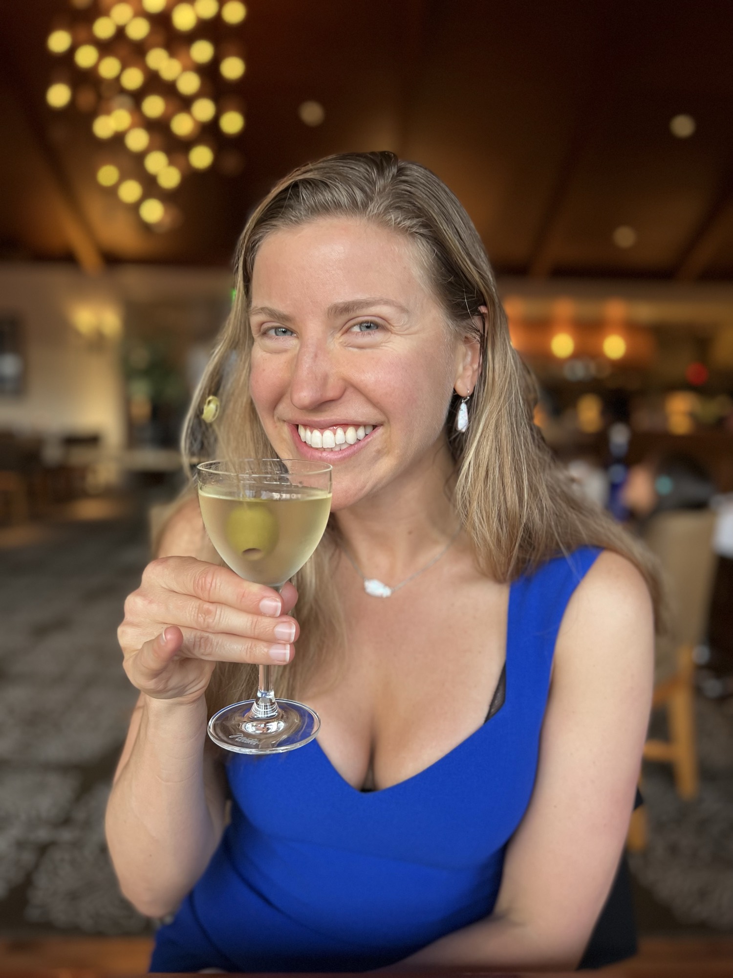 A smiling woman in a blue dress holds up a martini glass with an olive inside while sitting in a restaurant. The background is softly blurred, showing warm, ambient lighting and the interior of the dining establishment.