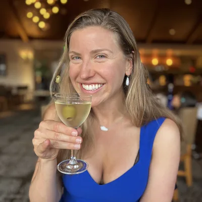 A smiling woman in a blue dress holds up a martini glass with an olive inside while sitting in a restaurant. The background is softly blurred, showing warm, ambient lighting and the interior of the dining establishment.