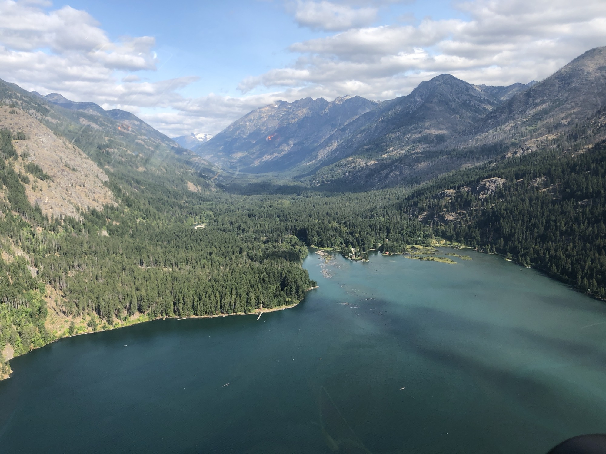 An aerial view captures a large, turquoise lake nestled in a valley surrounded by dense evergreen forests and rugged mountains under a partly cloudy sky. The perspective, likely taken from an airplane or helicopter, shows the vast wilderness landscape stretching toward distant peaks.