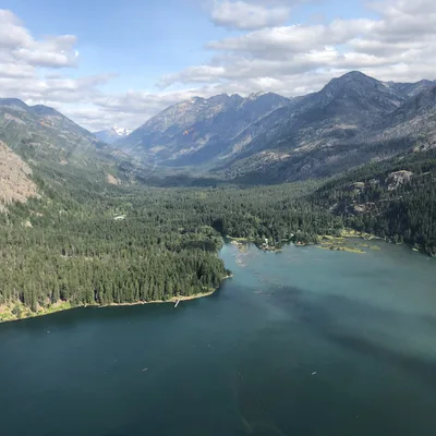 An aerial view captures a large, turquoise lake nestled in a valley surrounded by dense evergreen forests and rugged mountains under a partly cloudy sky. The perspective, likely taken from an airplane or helicopter, shows the vast wilderness landscape stretching toward distant peaks.